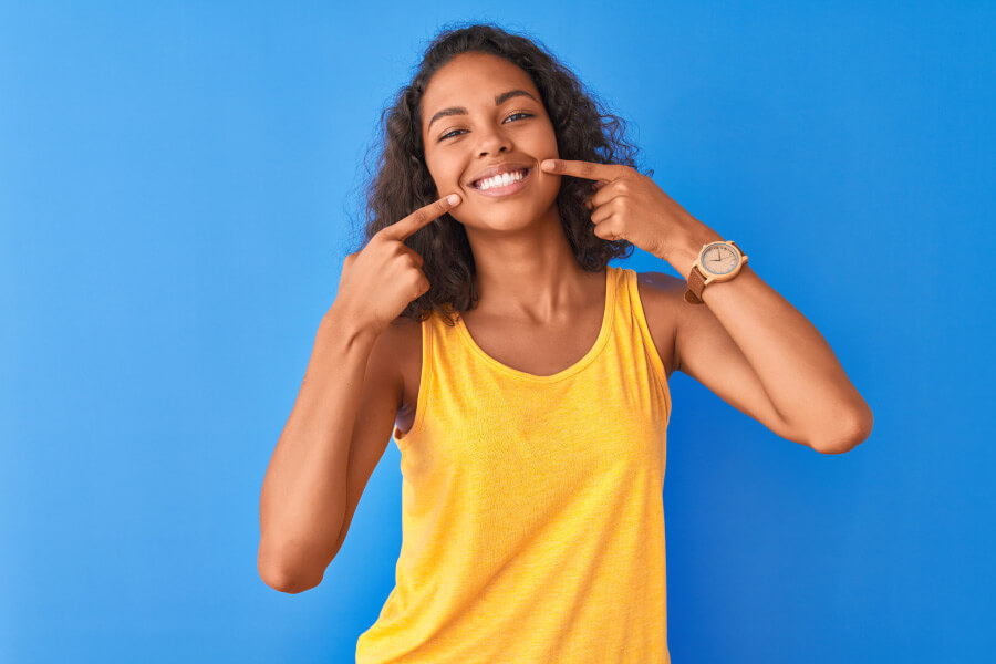 Brunette woman smiles in a yellow tanktop against a blue wall while pointing to her teeth